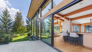 A modern kitchen with a bar area, large windows, and a view of the surrounding forest. - New luxury chalet with hot tub & media (Whistler)