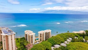 A coastal view with high-rise buildings, a clear blue ocean, and lush greenery. - Panoramic Oceanfront View, Breathtaking Sunsets, Relax on Large Lanai (Lahaina)