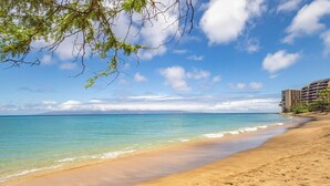 A beach with clear turquoise water, a sandy shore, and a building on the right. - Panoramic Oceanfront View, Breathtaking Sunsets, Relax on Large Lanai (Lahaina)