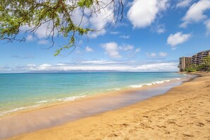 A beach with clear turquoise water, a sandy shore, and a building on the right. - Panoramic Oceanfront View, Breathtaking Sunsets, Relax on Large Lanai (Lahaina)