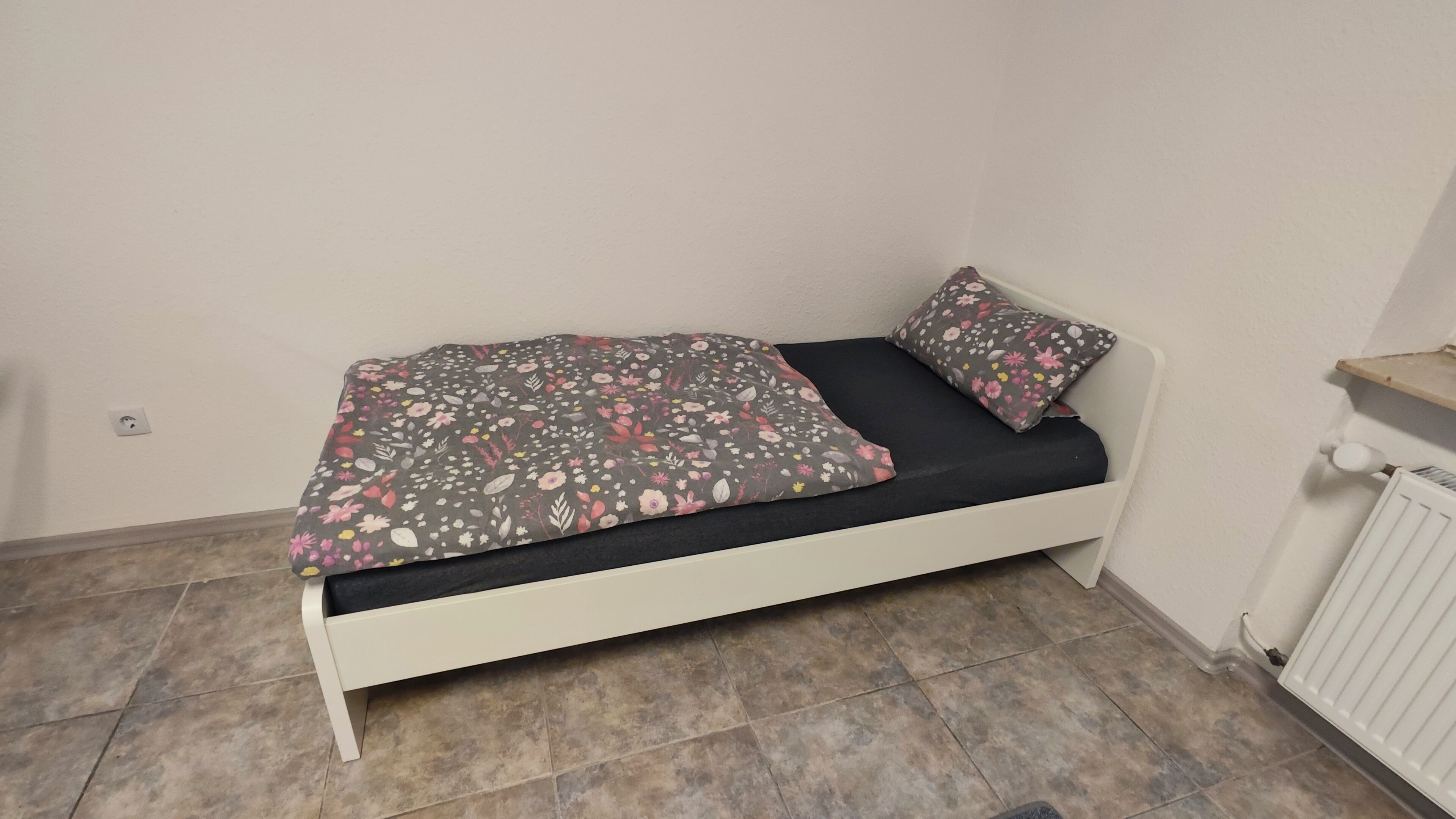 A small, single-bed room with a white frame, a dark mattress, and a floral-patterned bedspread.