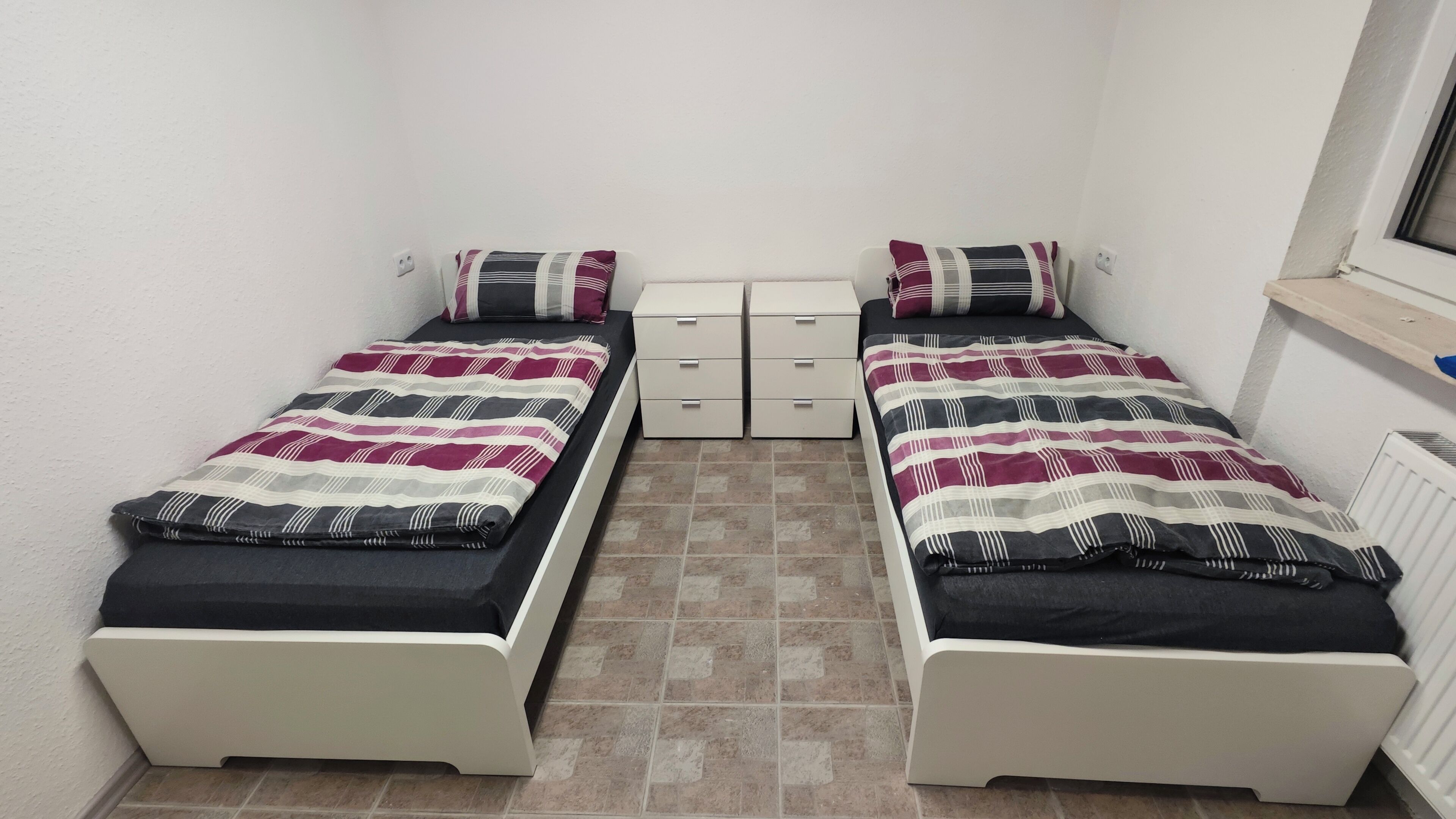 Une petite chambre propre, avec deux lits simples, une petite commode et une fenêtre.