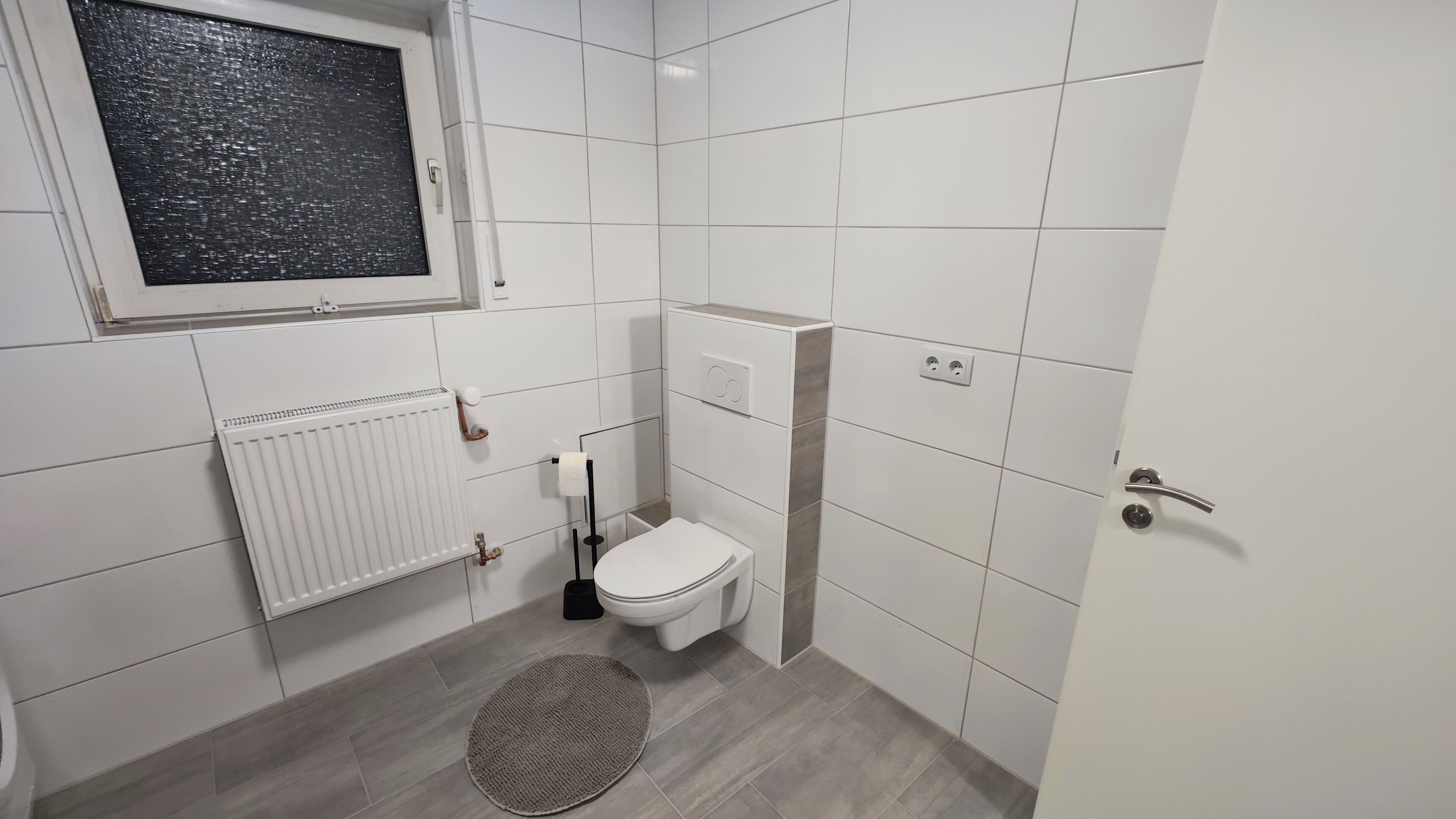 A small, modern bathroom with white tiles, a toilet, a radiator, a window, and a door.