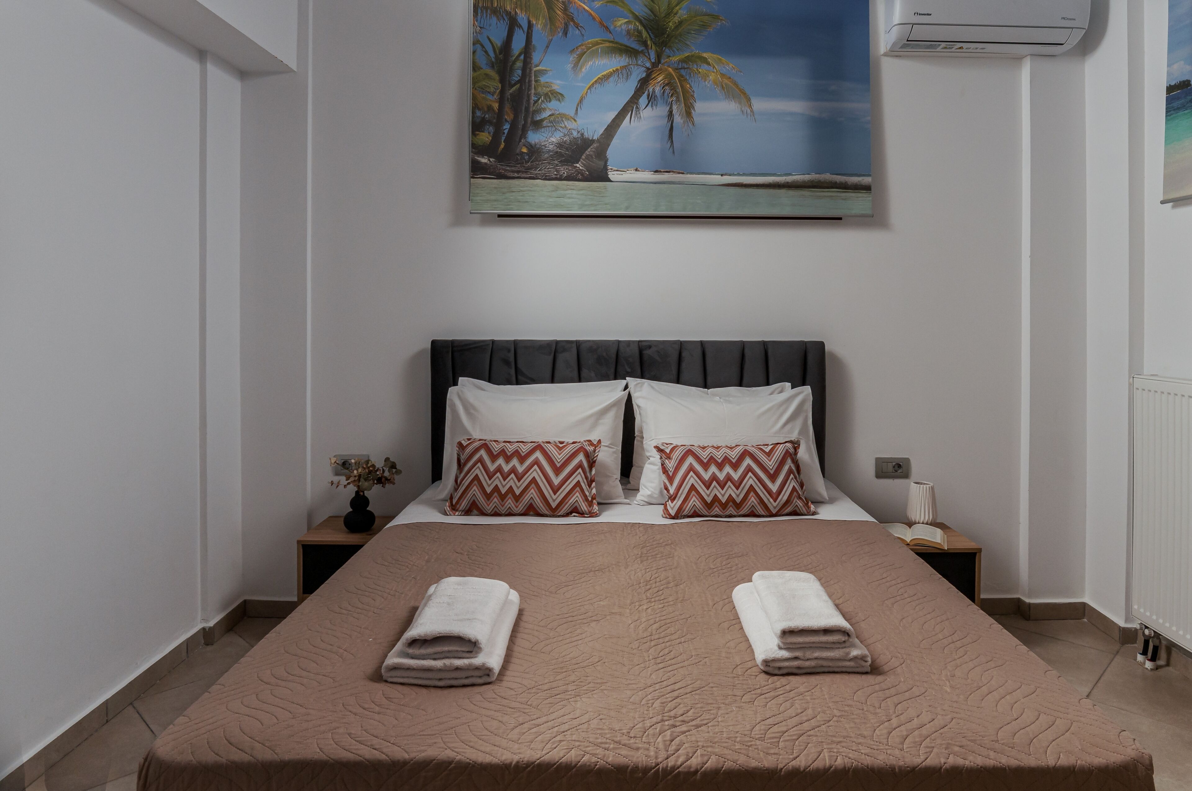 Un lit bien fait, recouvert d’une couverture marron et agrémenté de deux oreillers, une table de chevet avec une lampe et un livre, et un tableau représentant une scène de plage, accroché au-dessus du lit.