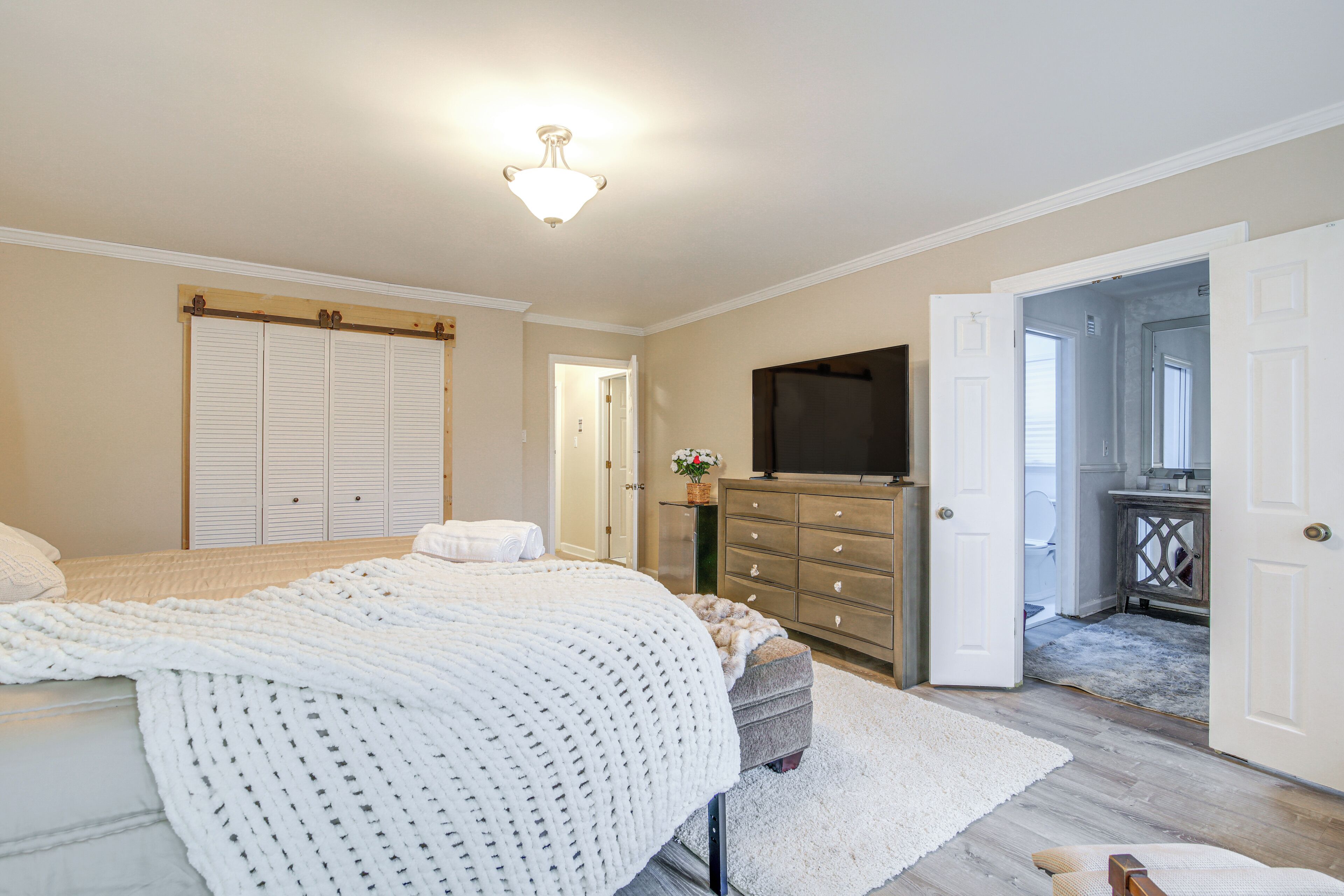 Un dormitorio con cama, cómoda, televisor y baño visible a través de una puerta abierta.