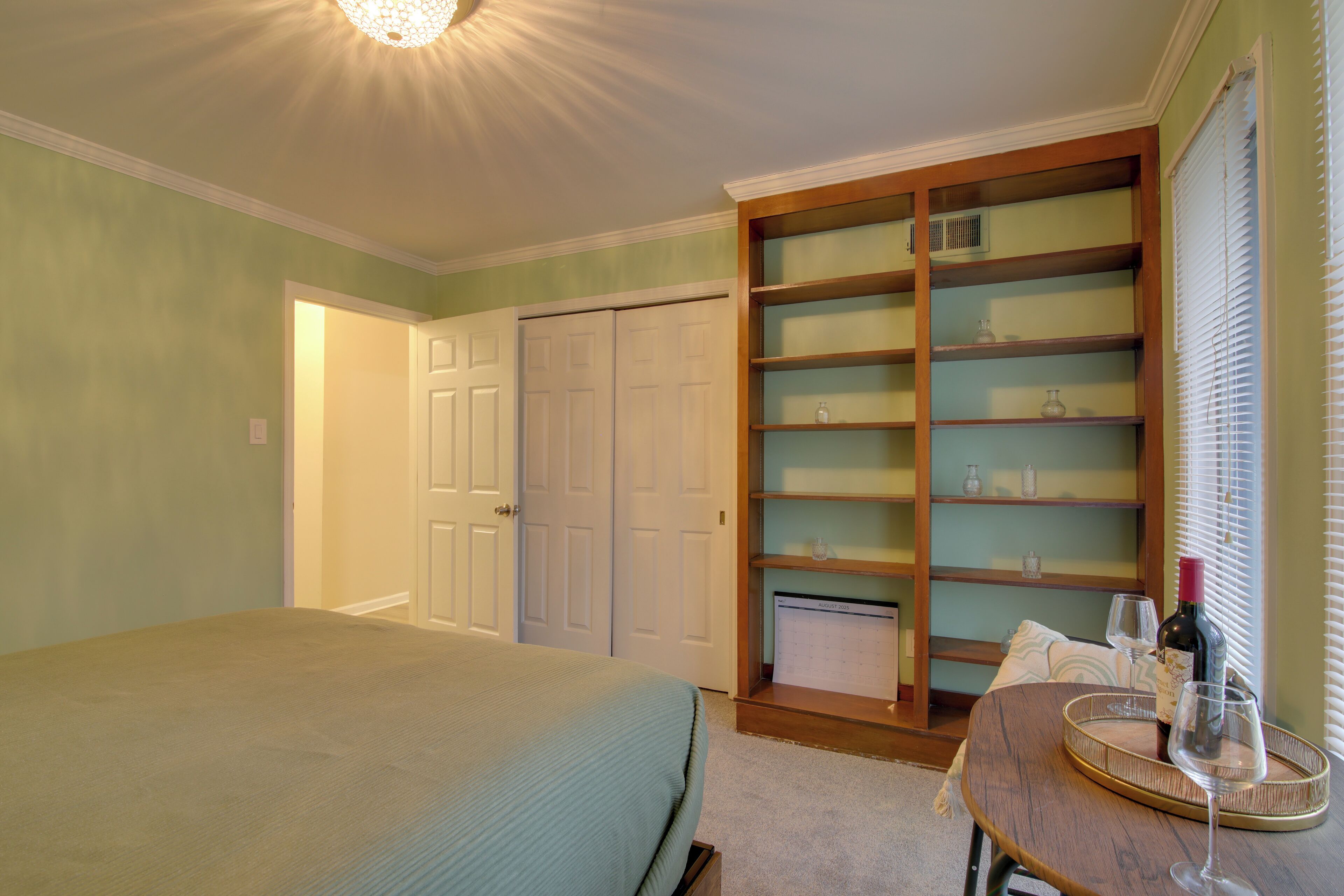 Un dormitorio con una cama, una mesita de noche de madera con una botella de vino y una copa, una estantería empotrada y una puerta que da a otra habitación.