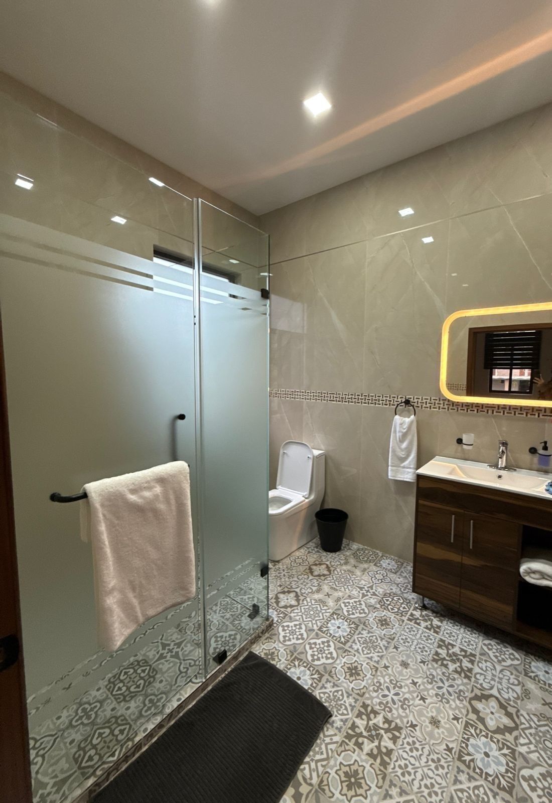 A modern bathroom with a glass shower enclosure, a toilet, a sink with a mirror, and patterned floor tiles.