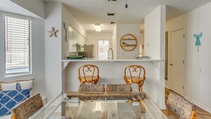 Un comedor con una mesa de vidrio rodeada por cuatro sillas de madera con cojines estampados, una pared blanca con un adorno de estrella y un estante de madera sobre el fregadero.