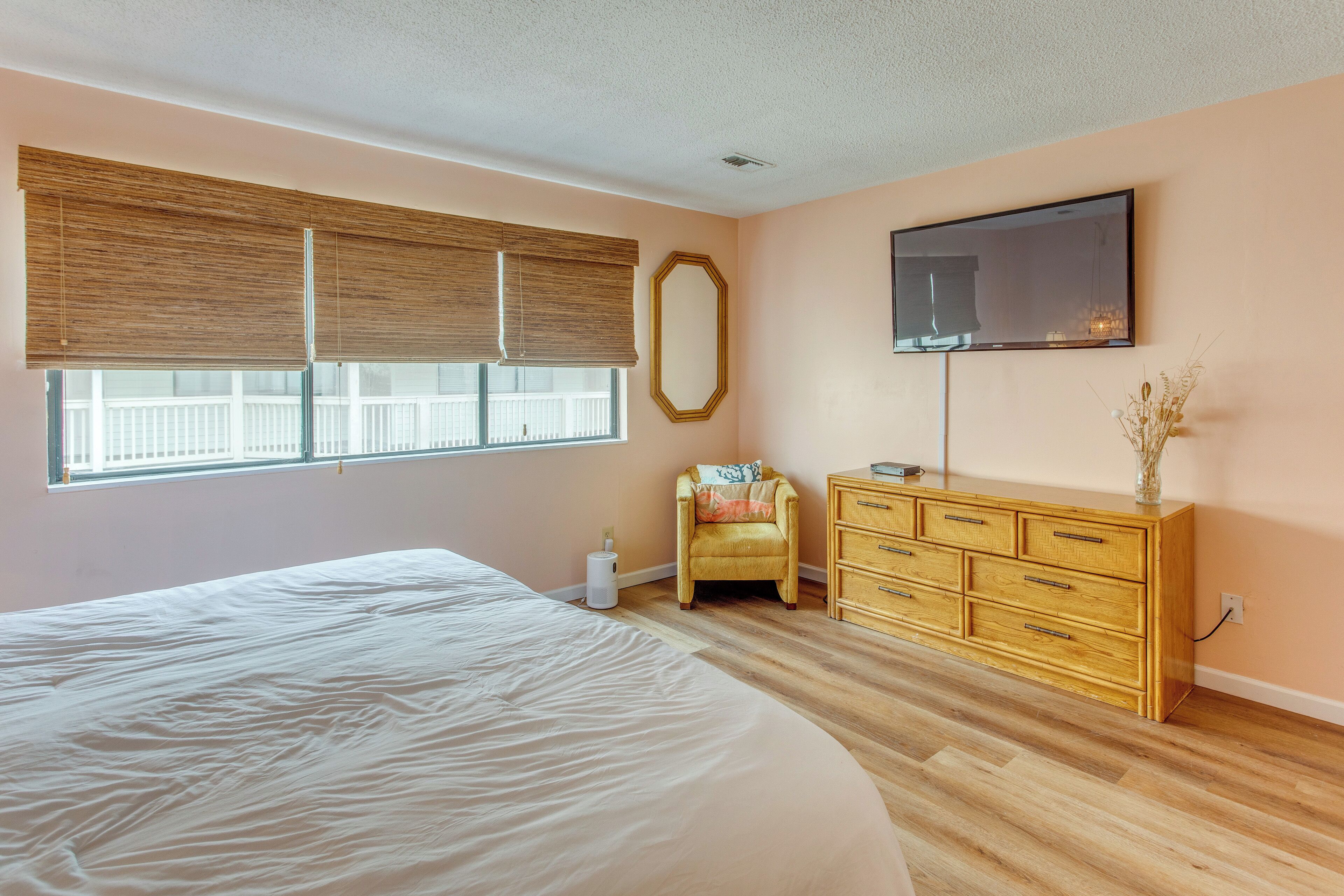 A bedroom with a large bed, a wooden dresser, a chair, a TV mounted on the wall, and a window with blinds.