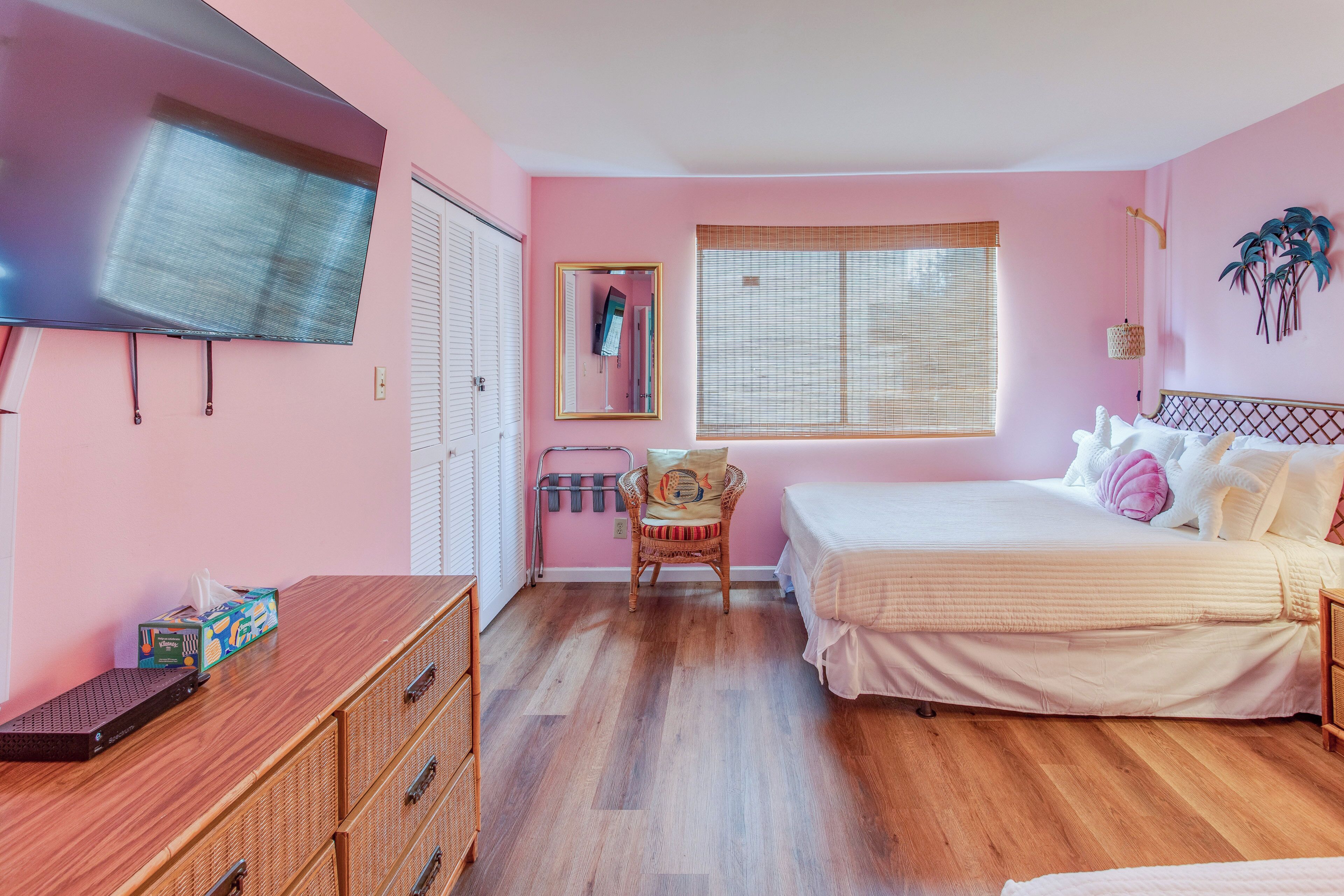 Un dormitorio con temática rosa, que incluye una cama, una silla, un aparador, un televisor y una ventana con persianas.