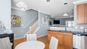 A modern kitchen with wooden cabinets, a stainless steel dishwasher, and a white dining area with a round table and cushioned chairs. - Rehoboth Crossing 1 - For 10 (Rehoboth Beach)