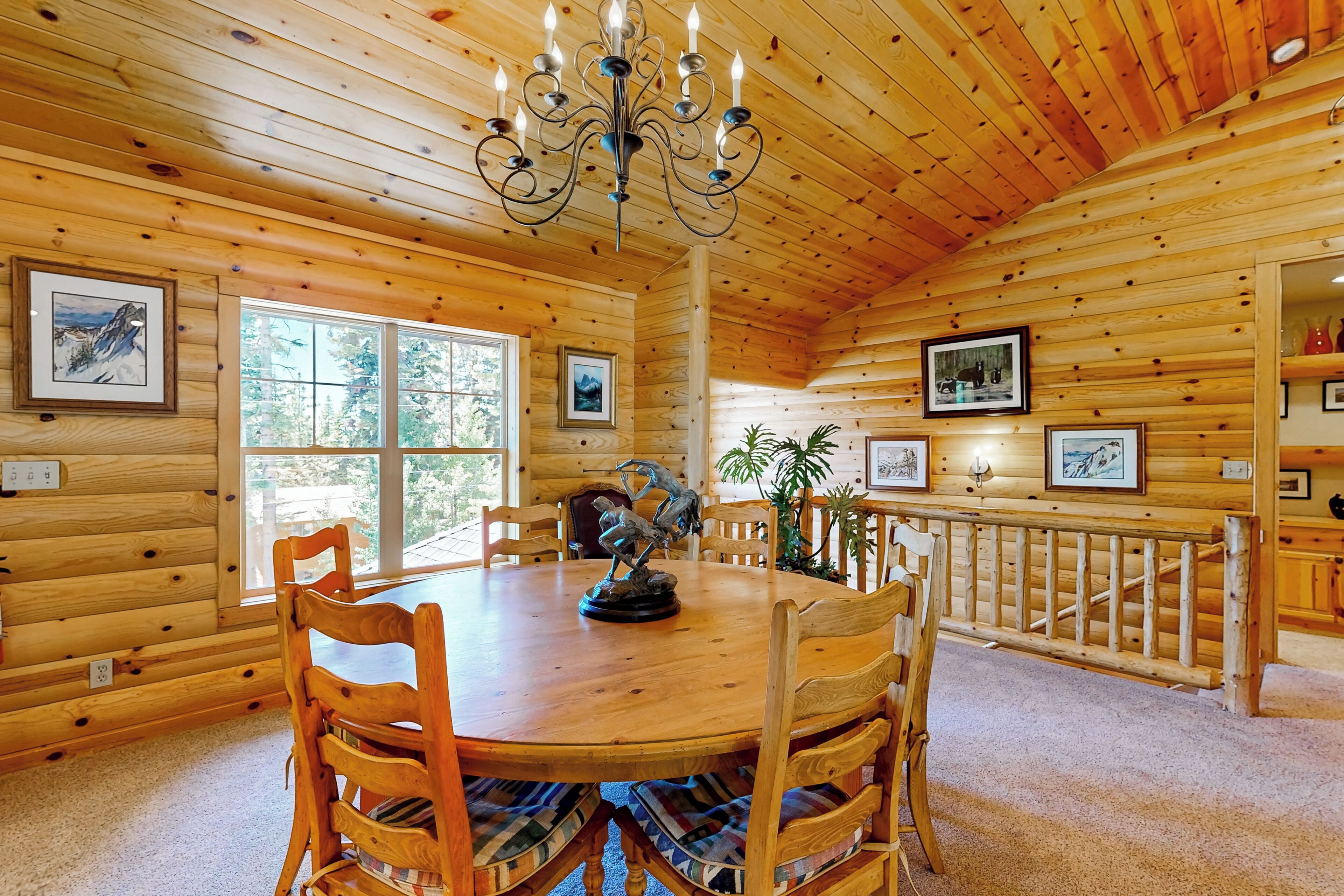 A wooden cabin interior with a round dining table, wooden chairs, a chandelier, framed pictures on the walls, and a window providing natural light.