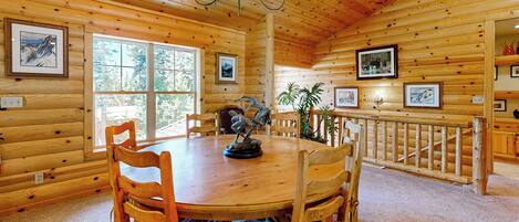 A wooden cabin interior with a round dining table, wooden chairs, a chandelier, framed pictures on the walls, and a window providing natural light.