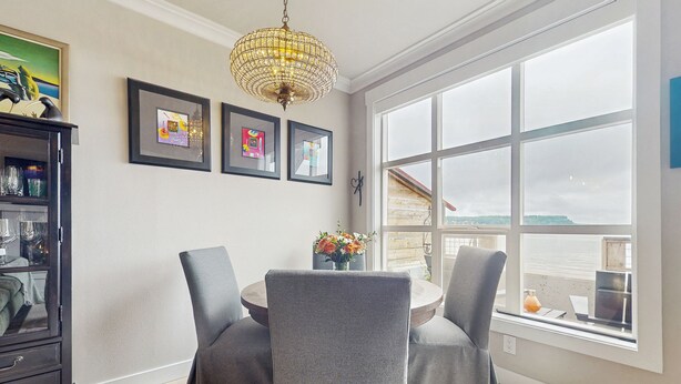 A dining area with a round table, two grey upholstered chairs, a chandelier, and framed artwork on the wall. - Studio Overlooking Saratoga Passage & Cascade Mtns (Langley)