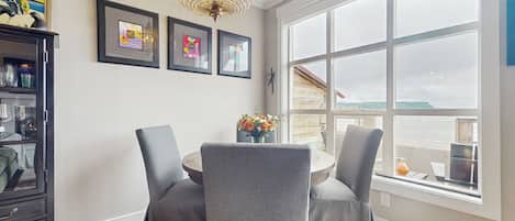 A dining area with a round table, two grey upholstered chairs, a chandelier, and framed artwork on the wall.
