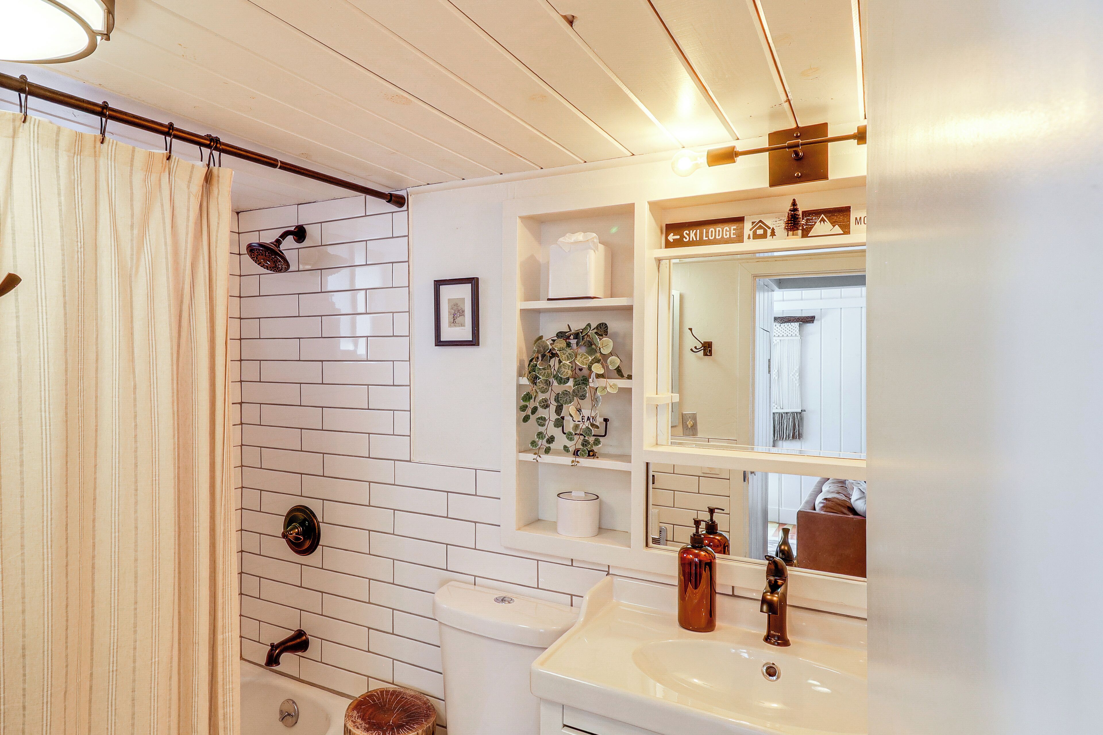 A bathroom with a shower, toilet, sink, and a mirror. There are shelves with toiletries and a framed picture on the wall.