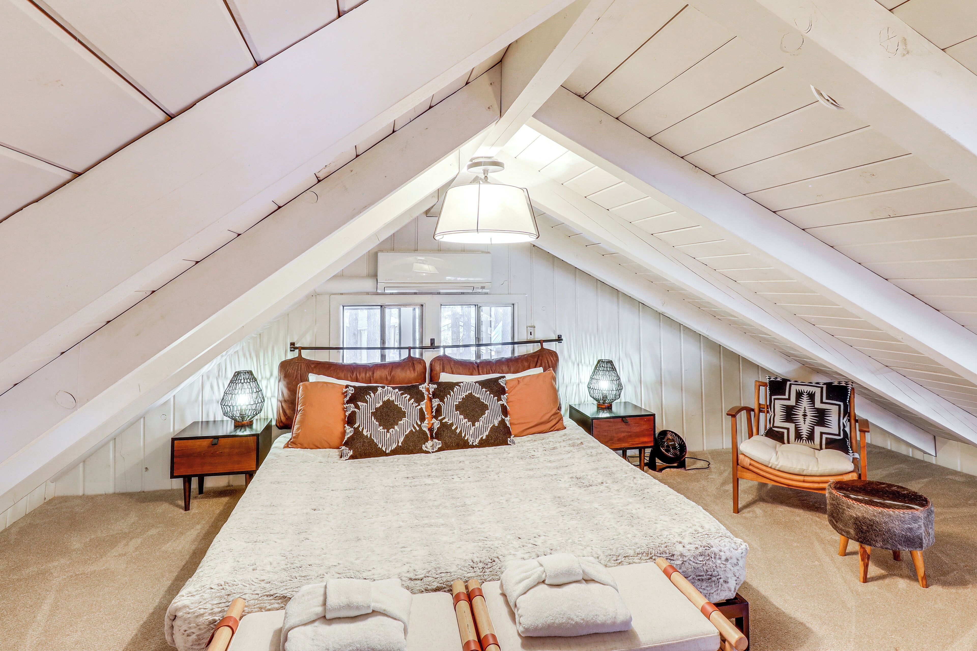 Un amplio dormitorio en el ático con una cama grande, vigas de madera, una lámpara colgante y vistas al exterior.