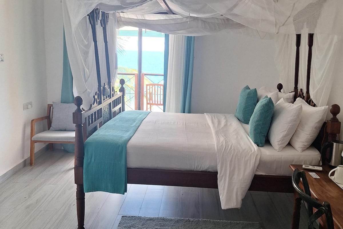A bedroom with a four-poster bed, a wooden chair, a small table, and a view of the ocean through a balcony.