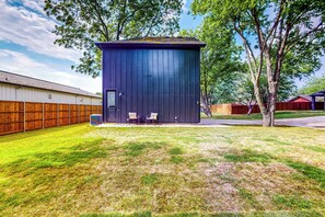 A small, dark-colored shed with a door and windows, surrounded by a wooden fence and a grassy yard. - 'mckinney Marvel' Studio in Historic District (McKinney)