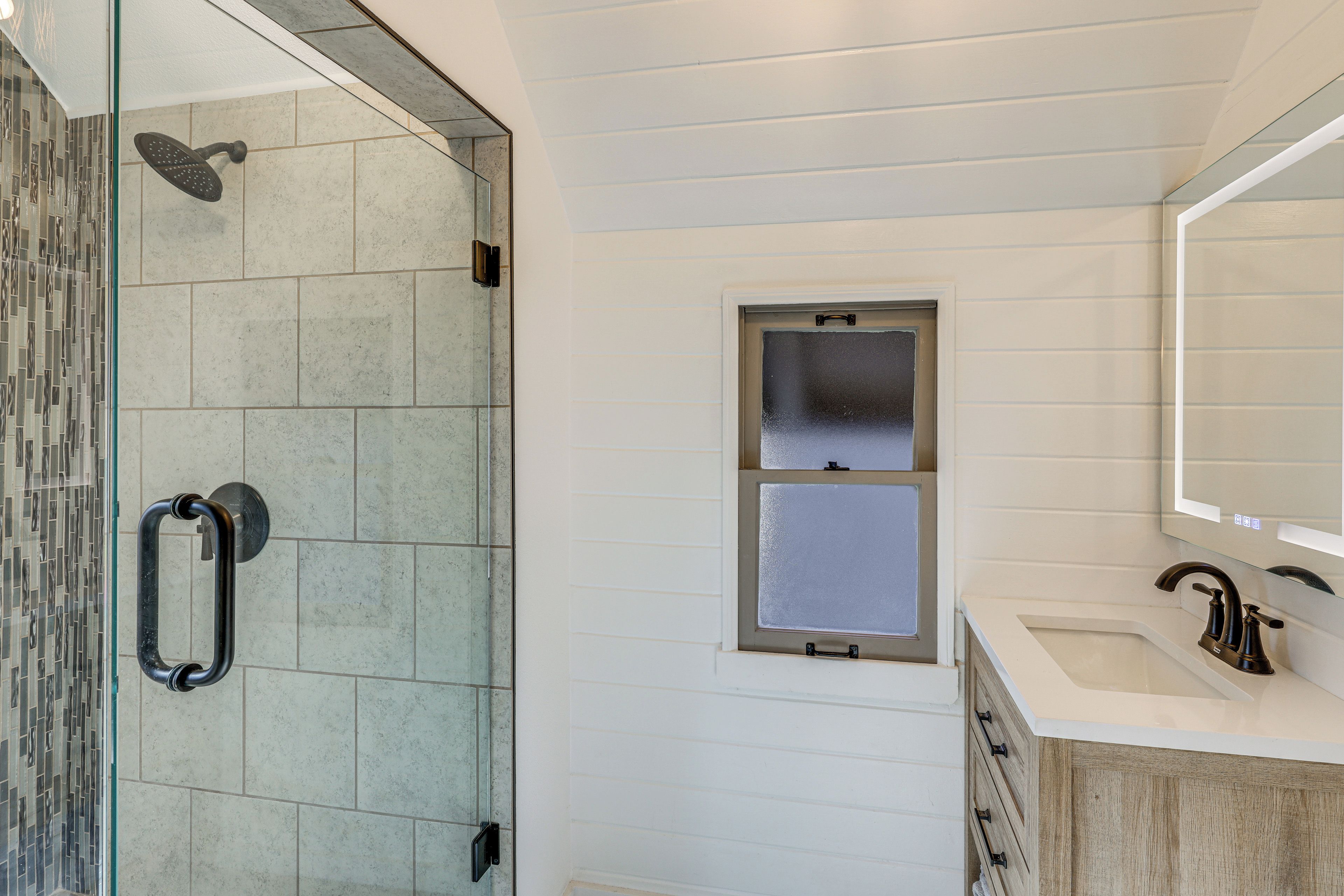 A bathroom with a glass-enclosed shower, a window, and a sink with a mirror above it.