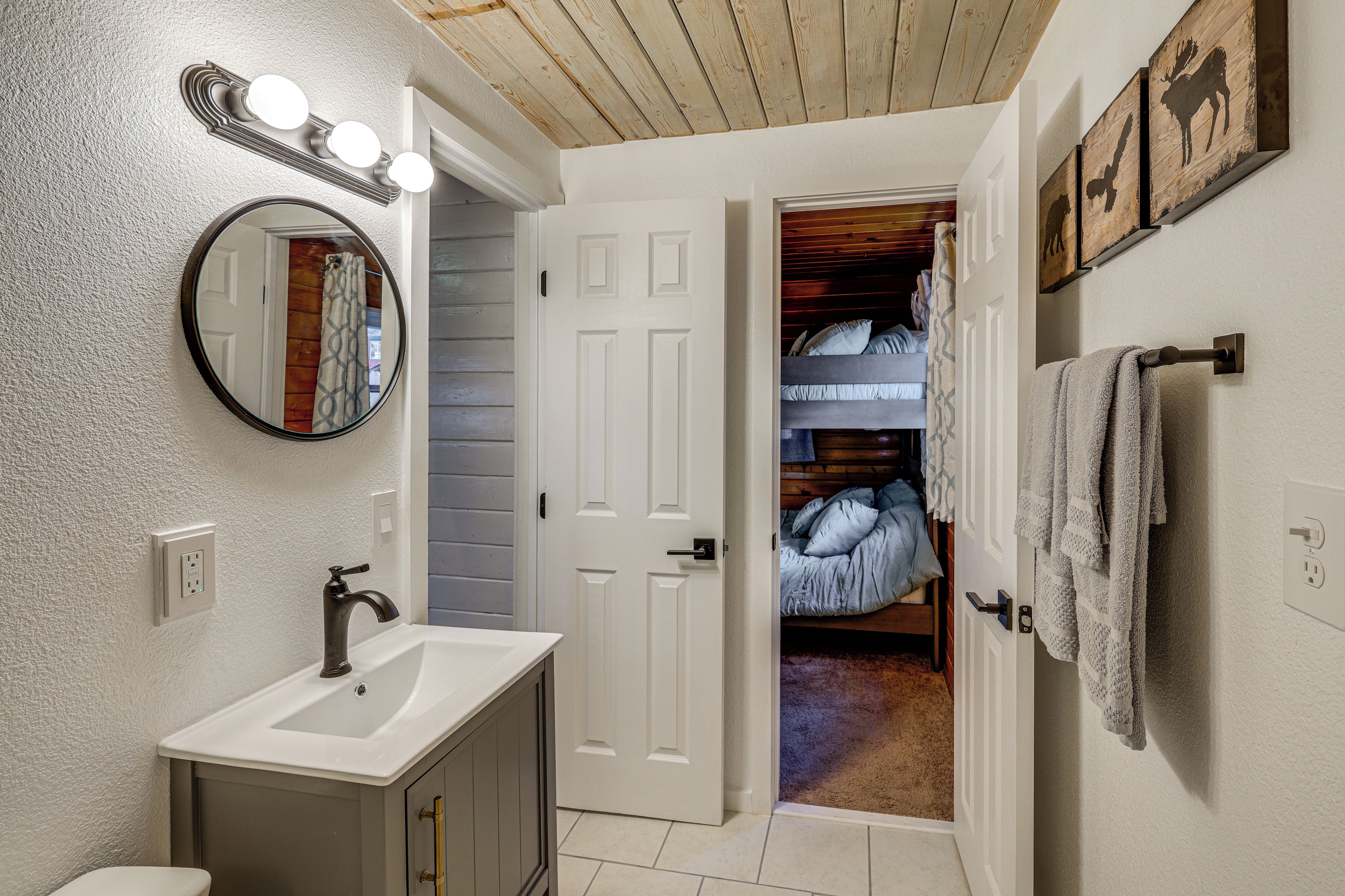Un baño con lavamanos, espejo y tocador. Hay una puerta que da a un dormitorio con cama y perchero.