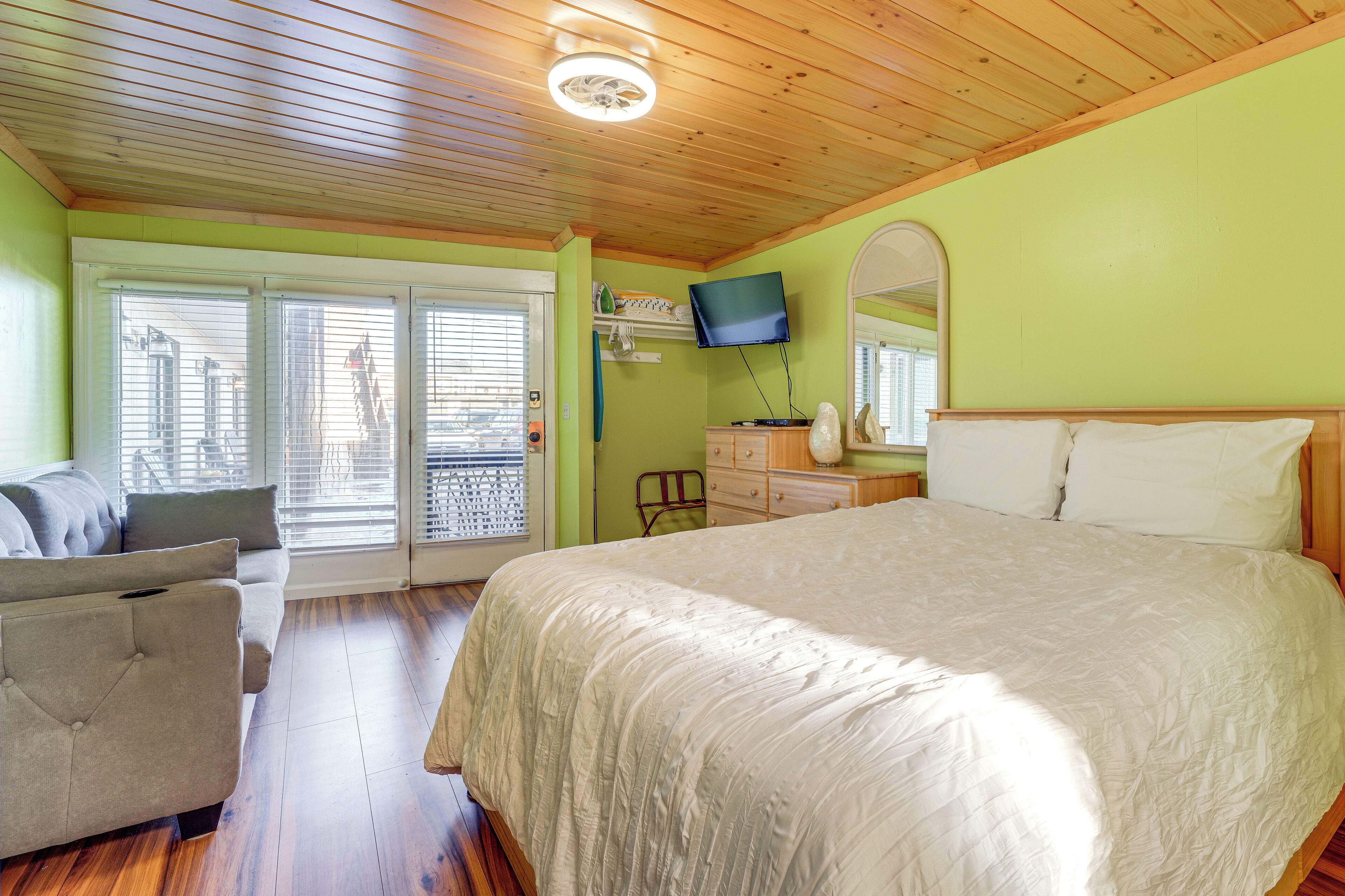 A bedroom with a large bed, a sofa, a television mounted on the wall, and a sliding glass door leading to an outdoor area.