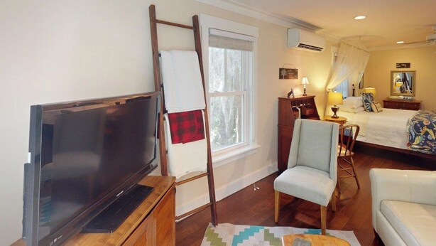 A hotel room with a flat-screen TV, a ladder leaning against the wall, a bed with a canopy, a chair, and a bedside table with lamps. - Steps to Riverfront: Dtwn Bluffton Studio! (Bluffton)