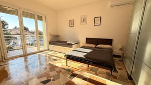 Ein Schlafzimmer mit Bett, einer Kommode, einem kleinen Tisch und einem Fenster mit Blick auf einen Balkon und Gebäude.
