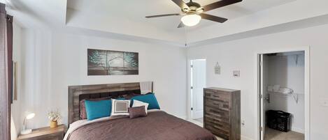 A bedroom with a large bed, a ceiling fan, a nightstand with a lamp, a wooden dresser, and a closet with shelves.
