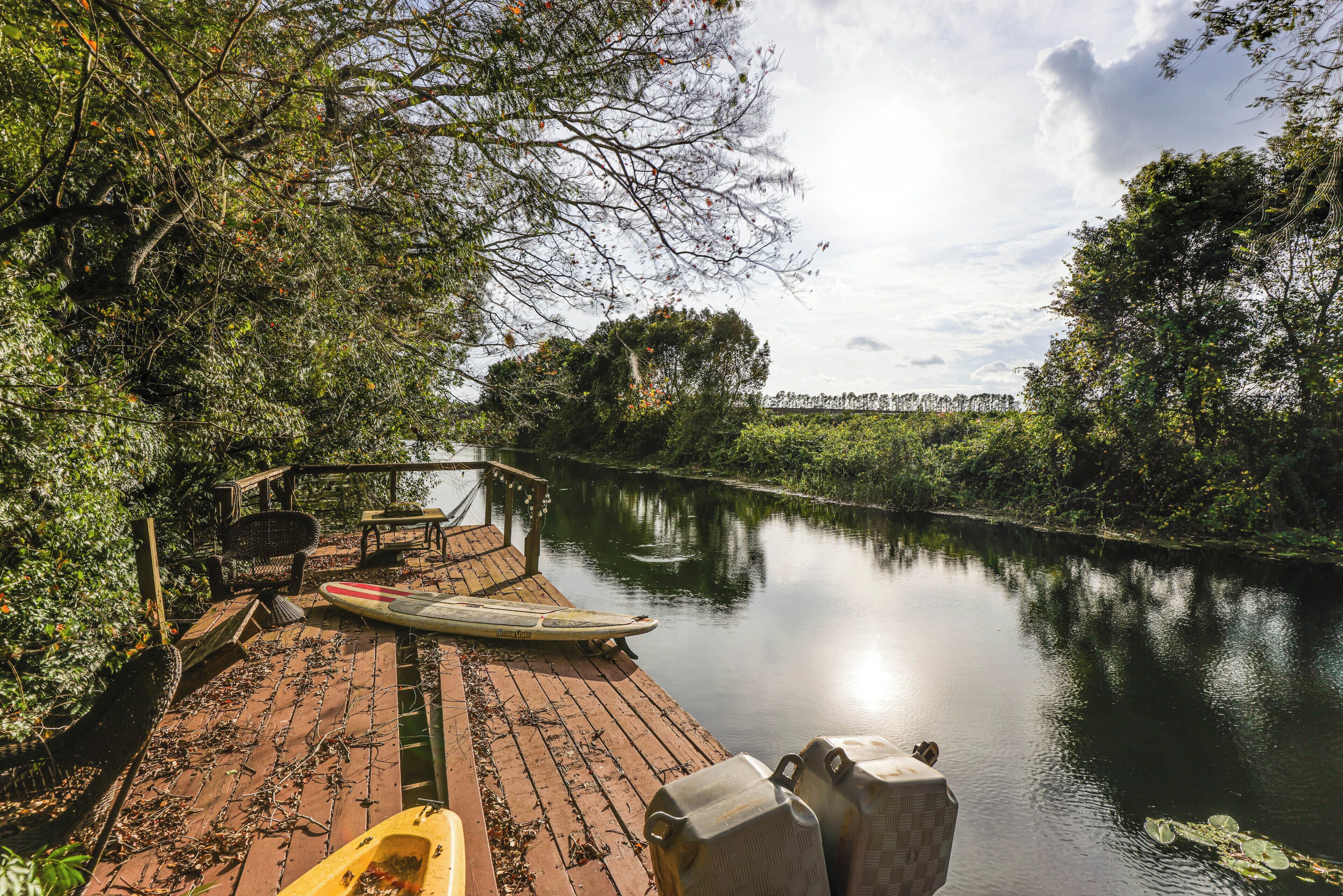 Une jetée en bois, avec un kayak et une planche à pagaie, entourée d’une végétation luxuriante et bordée par une rivière paisible.