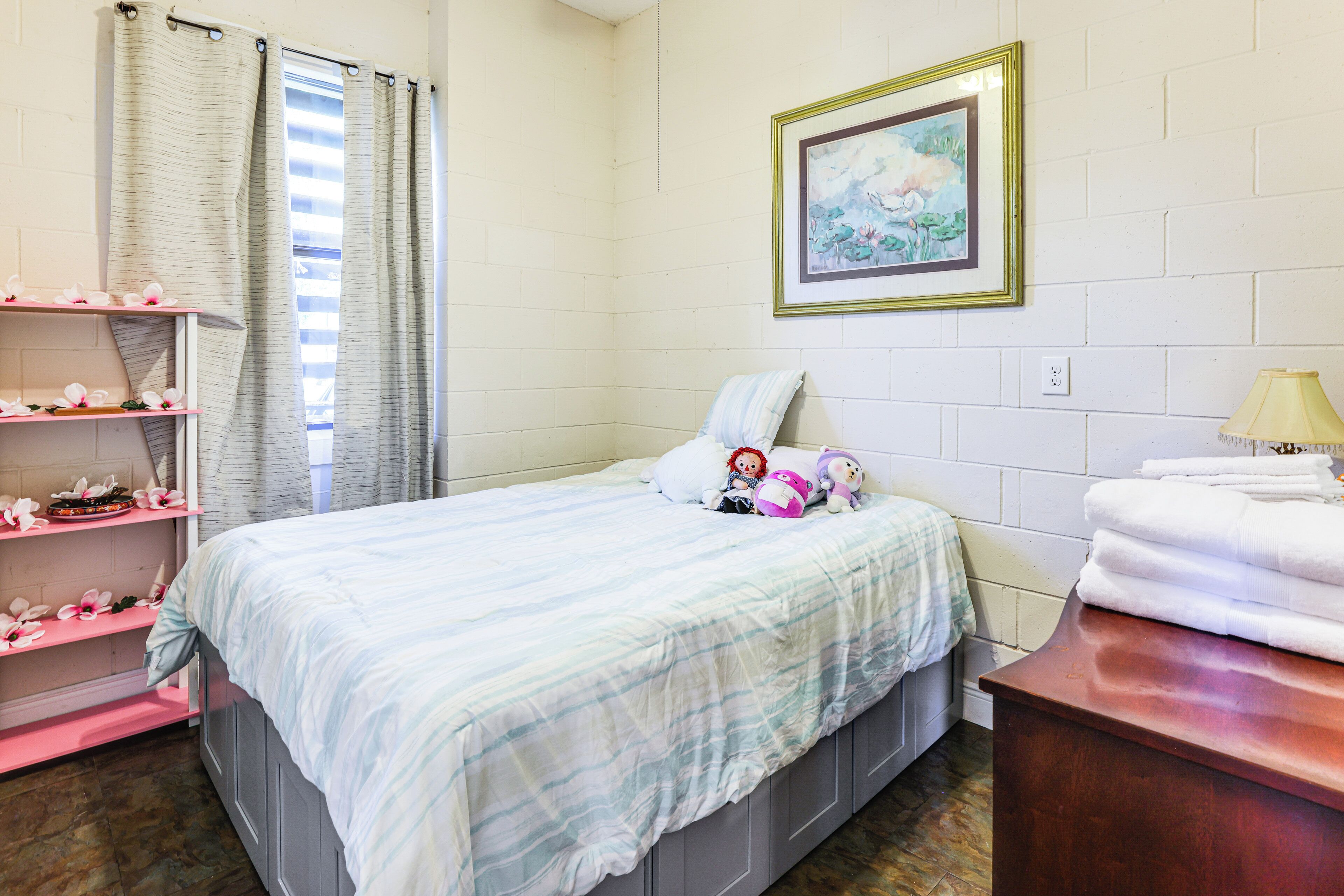 A bedroom with a bed, a shelf with decorative items, a framed picture on the wall, and a lamp on a bedside table.