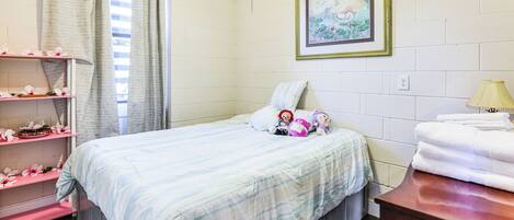 A bedroom with a bed, a shelf with decorative items, a framed picture on the wall, and a lamp on a bedside table.