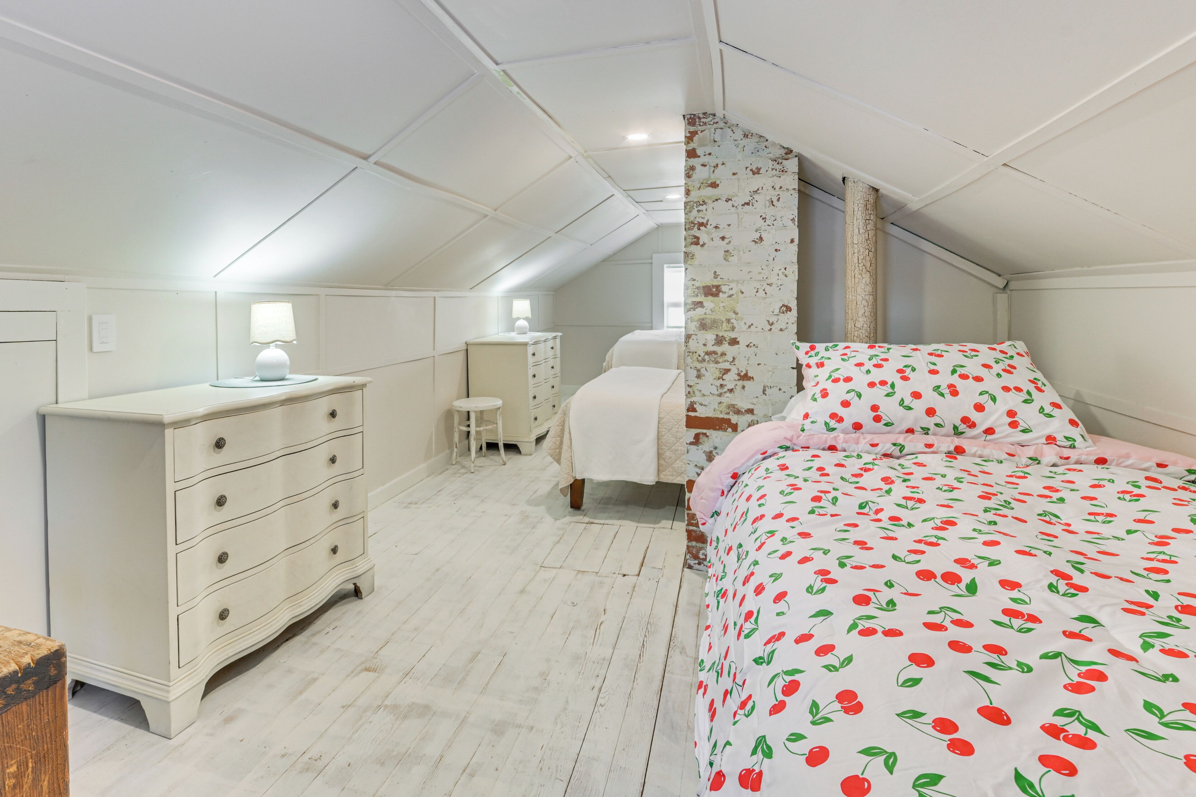 A bedroom with a sloped ceiling, a bed with a cherry pattern bedspread, a dresser, a nightstand, and a lamp.