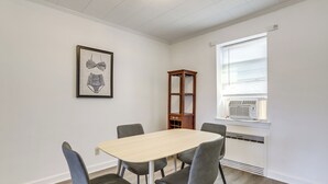 Una habitación con una mesa de madera y cuatro sillas, un cuadro enmarcado en la pared, un armario de madera y un equipo de aire acondicionado cerca de la ventana.