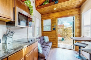 A compact kitchen with wooden cabinetry, a built-in microwave, and a countertop with a knife block. - Modern Tiny House in Hill Country w/ Covered Patio (Harper)