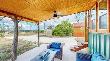 A covered outdoor area with a ceiling fan, two sofas with blue cushions, a table, and a staircase leading to a wooden building.