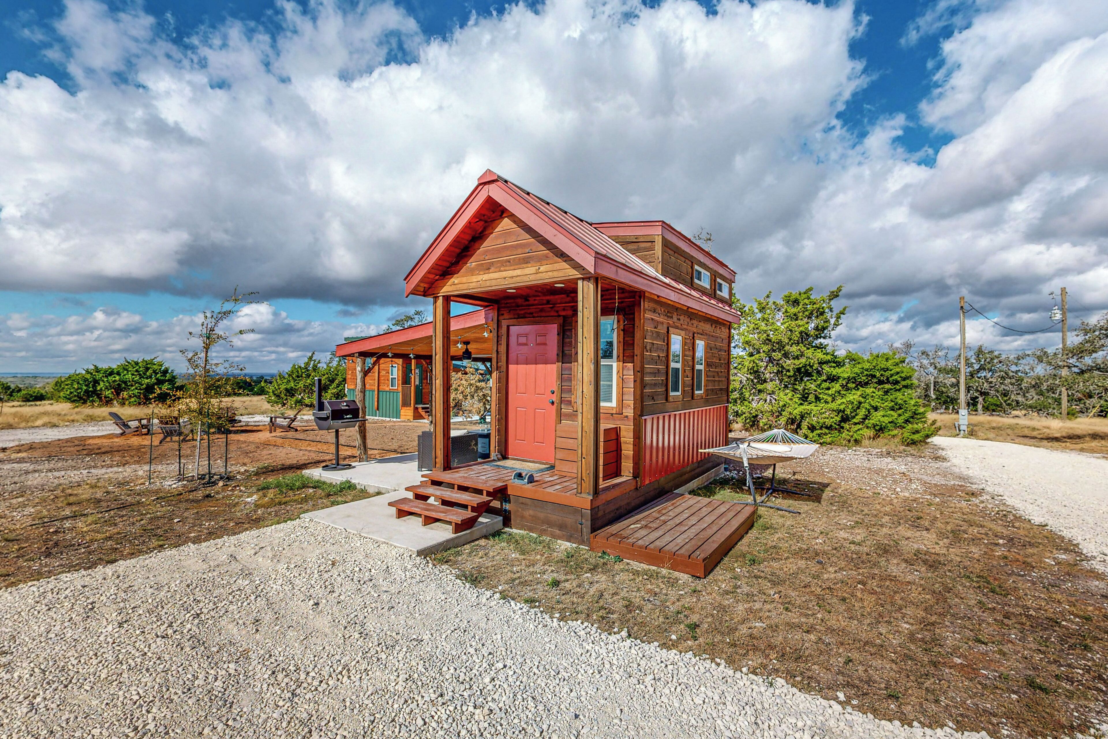 A small wooden cabin with a porch, a red door, and a gravel driveway.