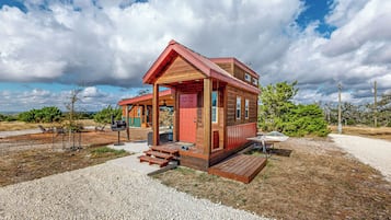 A small wooden cabin with a porch, a red door, and a gravel driveway.