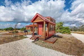 A small wooden cabin with a porch, a red door, and a gravel driveway.