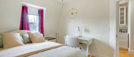 Ein Schlafzimmer mit Bett, Schreibtisch, Stuhl, einer Uhr und einem Fenster mit Vorhängen.