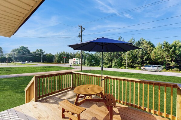 A wooden deck with a table and chairs, a blue umbrella, and a view of a road and trees. - Dog-friendly A-frame Cabin w/ Deck (Leesville)