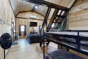 Innenansicht einer Holzhütte mit einem Etagenbett, einem Ventilator, einer Couch und einer Treppe.