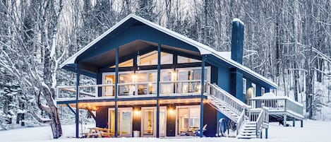 A two-story lodge with a balcony and a staircase, surrounded by snow-covered trees.