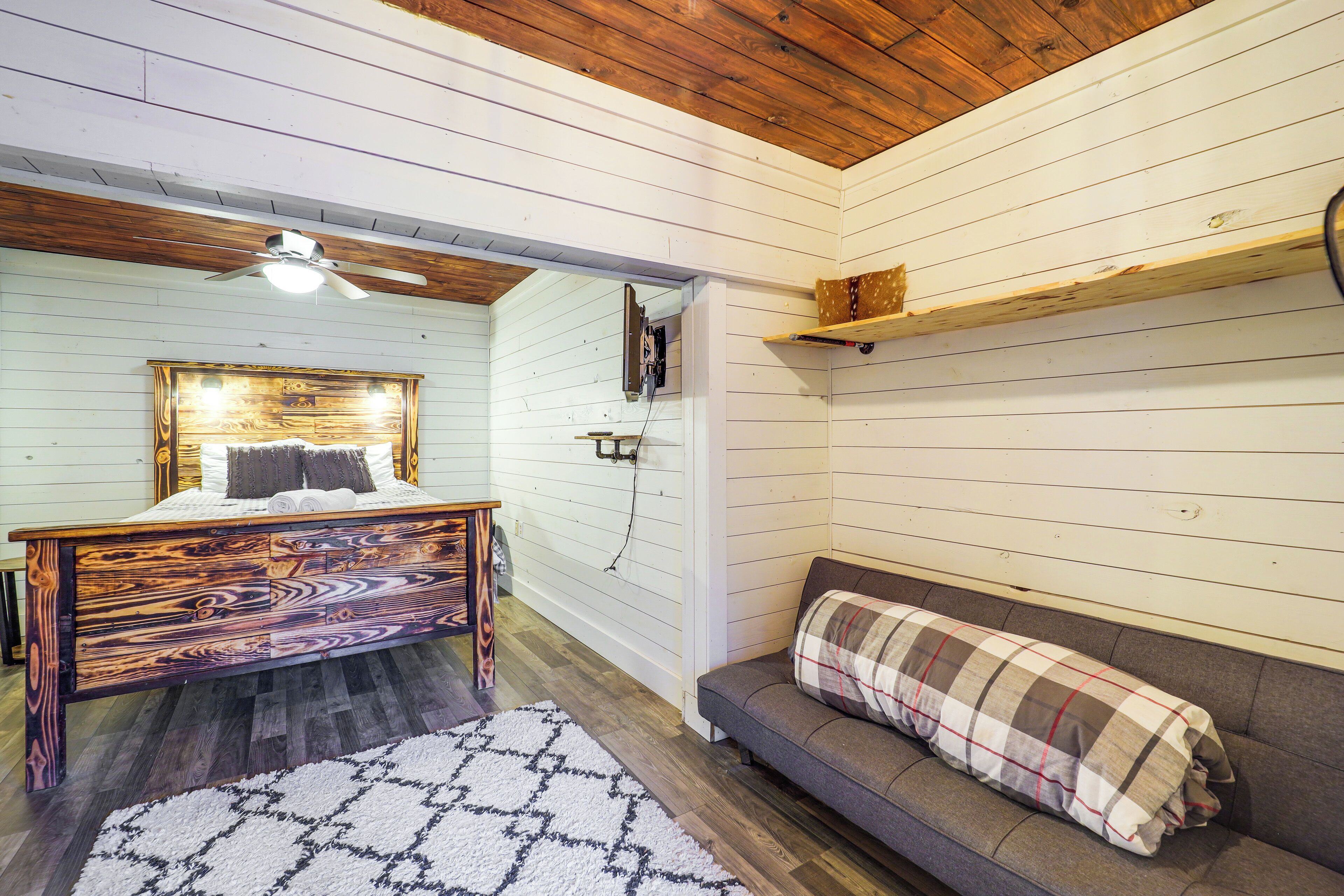 Une chambre à coucher avec un lit en bois, un canapé gris, un ventilateur de plafond et un mur blanc avec une étagère.