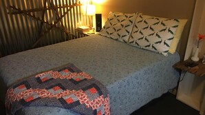Uma cama com colcha estampada, almofadas com padrão de folha, abajur e uma pequena mesa de cabeceira de madeira com um telefone e um objeto decorativo.