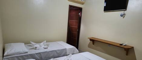 Une chambre d’hôtel avec deux lits, chacun recouvert d’une courtepointe blanche et agrémenté de serviettes disposées en motif floral. Un étagère en bois est fixée au mur, et un appareil de climatisation est installé au-dessus de la porte.