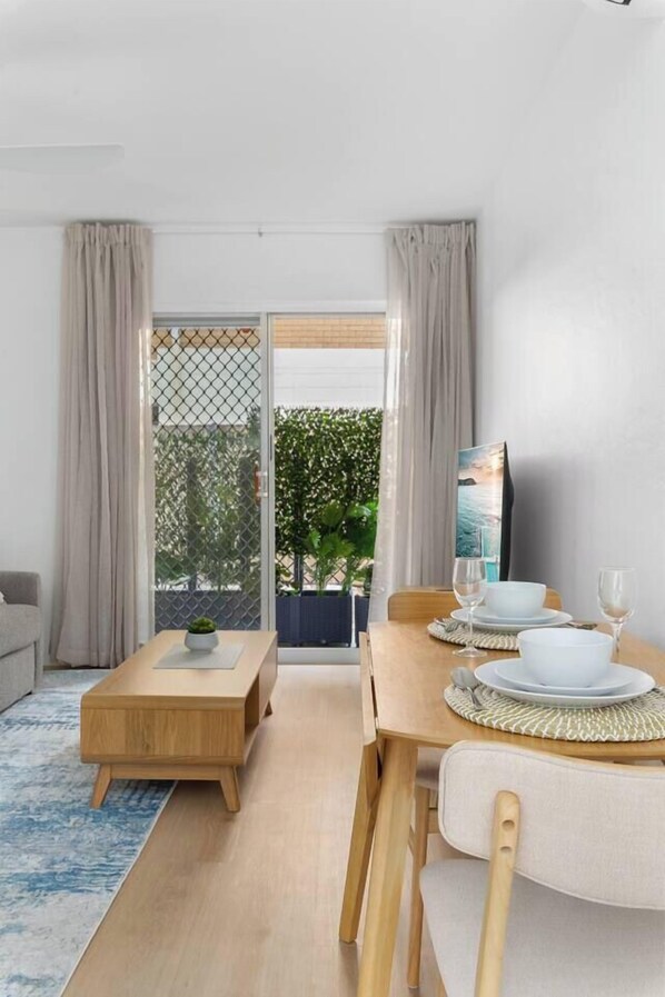 A dining area with a wooden table set for two, a small side table with a plant, and a view of greenery through a glass door.