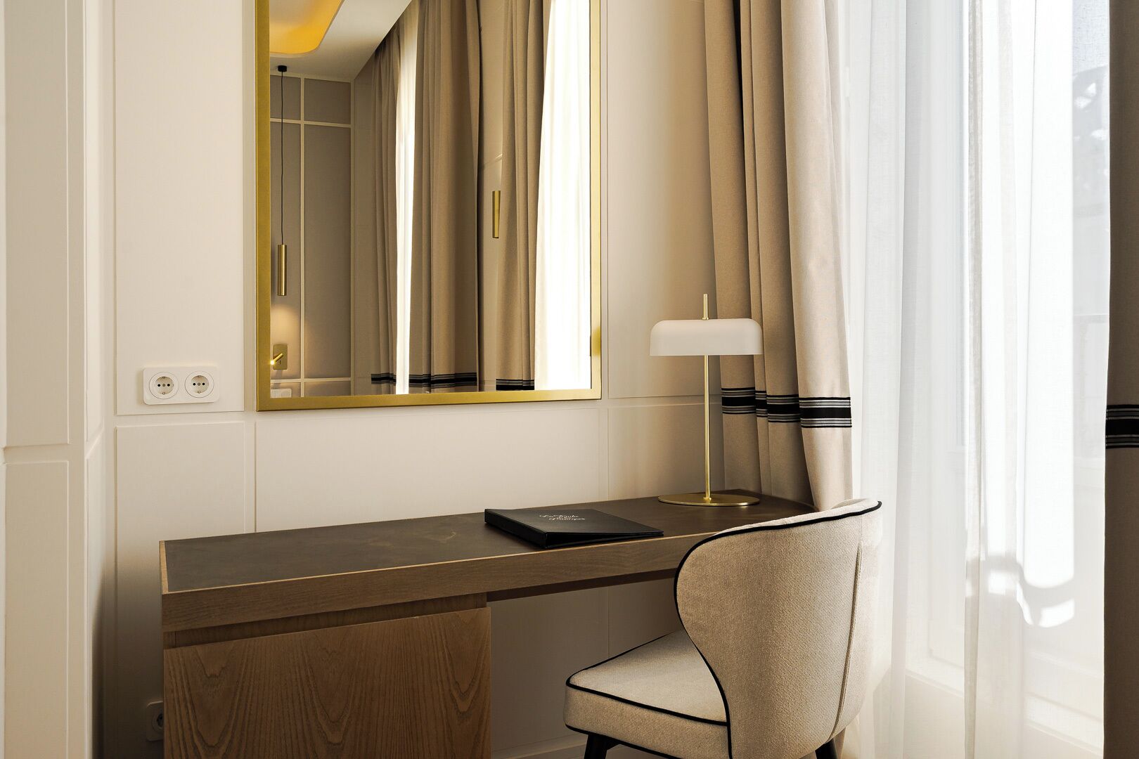 Habitación de hotel con escritorio de madera, una silla, lámpara, espejo y ventana con cortinas.