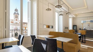 Een moderne hotel eetruimte met tafels en stoelen, grote ramen met uitzicht op een historisch gebouw, en hanglampen.