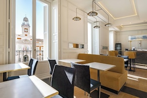 Area ristorazione di un hotel moderno con tavoli e sedie, ampie finestre con vista su un edificio storico e lampadari a sospensione.
